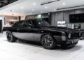 A shiny black classic muscle car, celebrated among high-performance cars, is displayed indoors on a white tile floor, surrounded by contemporary furniture and equipment.