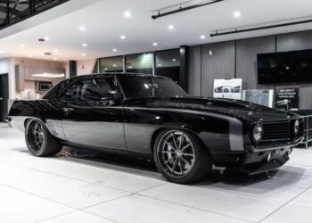 A shiny black classic muscle car, celebrated among high-performance cars, is displayed indoors on a white tile floor, surrounded by contemporary furniture and equipment.