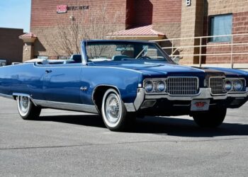A blue classic convertible car with whitewall tires, a true gem among Classic Muscle Cars, parked in front of a brick building on a sunny day.