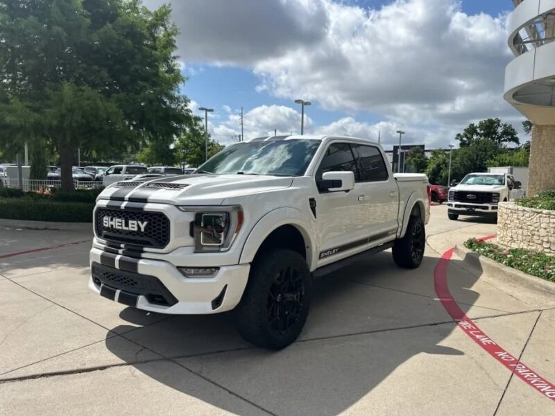 A white Shelby F-150 pickup truck with black racing stripes is parked on a paved lot near a curb under a partly cloudy sky, showcasing why Shelby trucks are so sought-after.