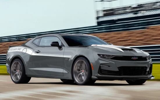 A gray Chevrolet Camaro muscle car driving on a racetrack with blurred background, featuring white racing stripes and sporty design elements.