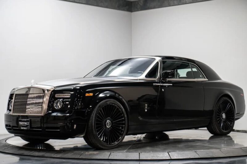 A black Rolls-Royce Phantom coupe is parked indoors on a circular display platform, photographed in a well-lit showroom—the type of luxury vehicle often favored by celebrity owners.