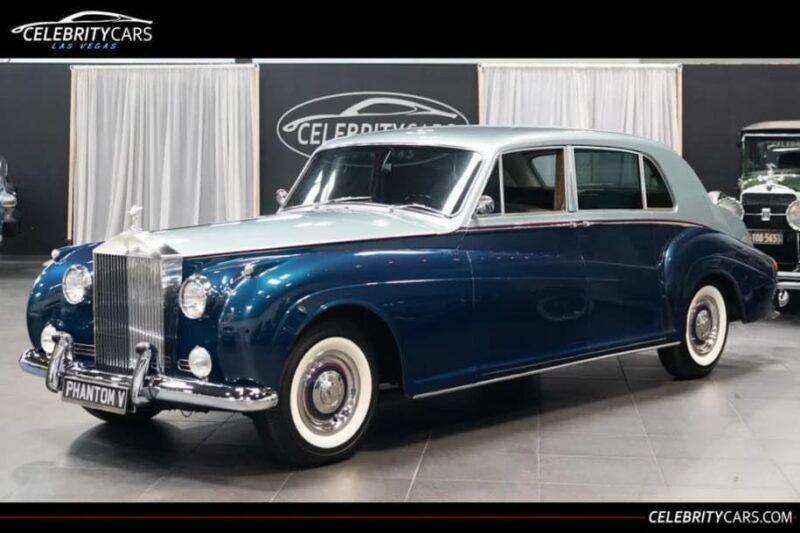 A classic blue and silver Rolls-Royce Phantom V sedan, favored by celebrity owners, is displayed indoors under showroom lighting.