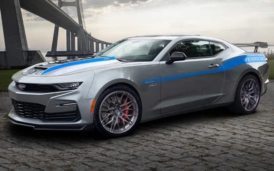 A silver Chevrolet Camaro muscle car with blue racing stripes is parked on a cobblestone surface near a large bridge.
