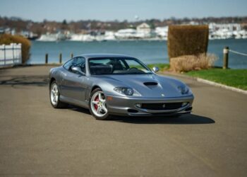 A silver Ferrari 550 Maranello, a stunning luxury car, is parked on a paved road near a body of water, with trees, grass, and buildings in the background.