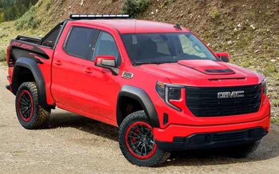 A red GMC off-road pickup truck with black accents and raised suspension, parked on a gravel road near a grassy slope—a rugged choice among trucks for sale from Specialty Vehicle Engineering.