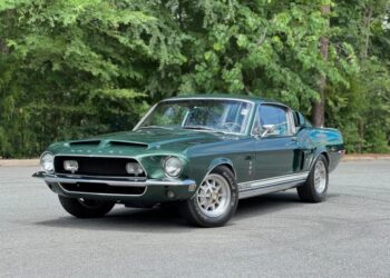 A dark green classic Ford Mustang Shelby GT350, one of the iconic Classic Muscle Cars, is parked on a paved surface, with trees and greenery in the background.