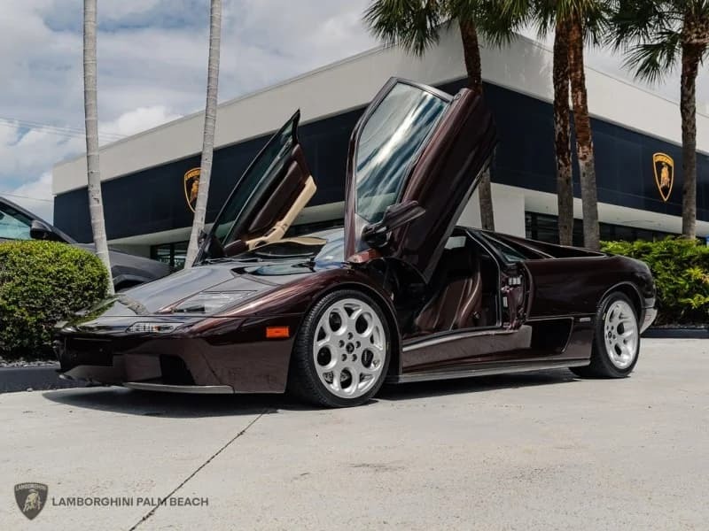 A dark-colored Lamborghini Diablo, an iconic exotic car with its scissor doors open, is parked outside a Lamborghini dealership surrounded by palm trees.