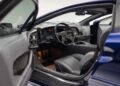 A view of a Jaguar XJ220 supercar’s interior through the open driver’s side door, showing the steering wheel, dashboard, and front seats in gray upholstery.