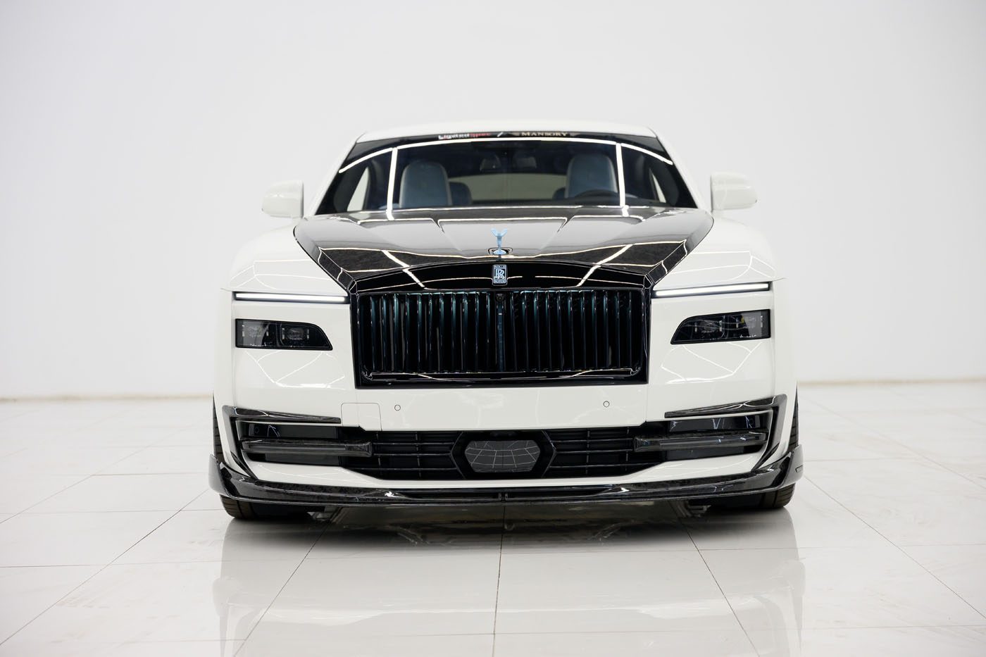 An exclusive 2025 Cullinan Mansory, featuring a striking black grille and hood, is parked indoors on a glossy white tiled floor, captured from the front.