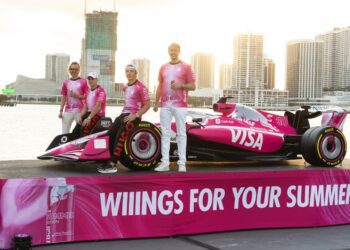 Four people in pink shirts stand beside a pink Formula 1 car with “VISA” branding, displayed on a platform with “WIIINGS FOR YOUR SUMMER” text, cityscape and water in background.