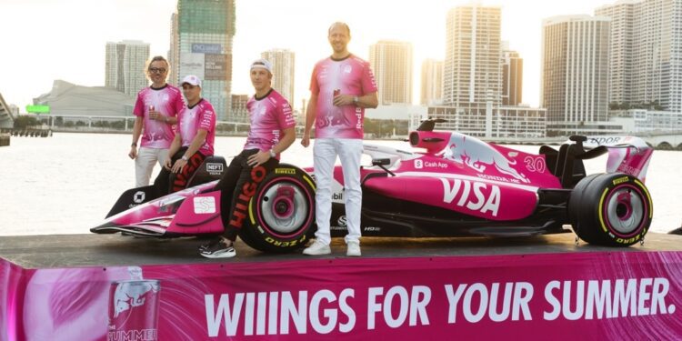 Four people in pink shirts stand beside a pink Formula 1 car with “VISA” branding, displayed on a platform with “WIIINGS FOR YOUR SUMMER” text, cityscape and water in background.