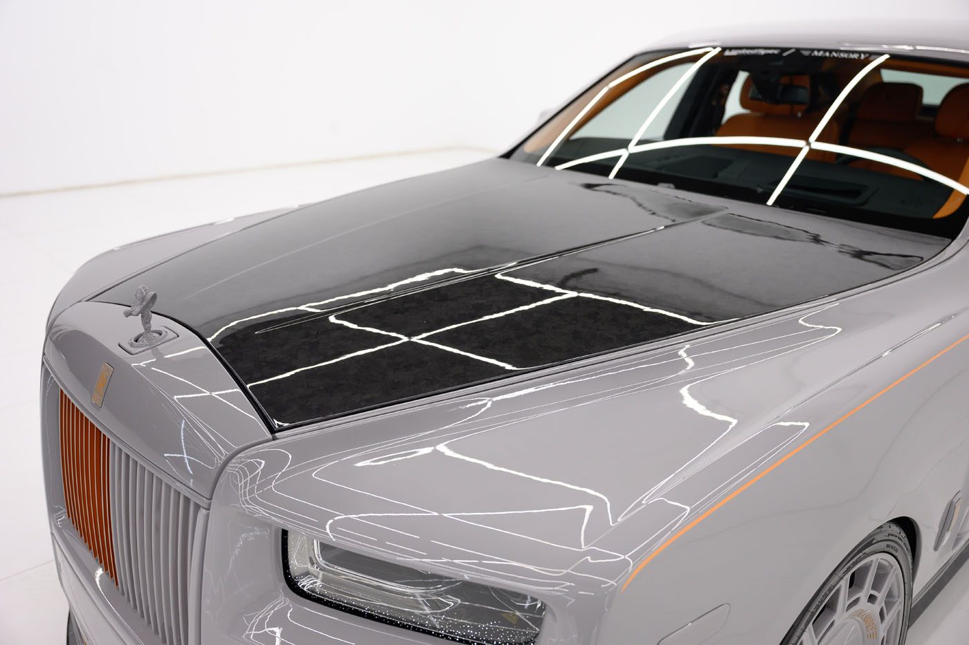 A close-up of the exclusive 2025 Cullinan with an exposed carbon fiber hood, orange accents, and a Spirit of Ecstasy hood ornament on display at a Mansory Dealer in a well-lit showroom.