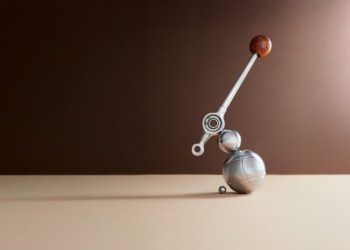 A metallic abstract sculpture with spheres and a lever-like arm topped with a wooden ball, placed on a beige surface against a brown gradient background.