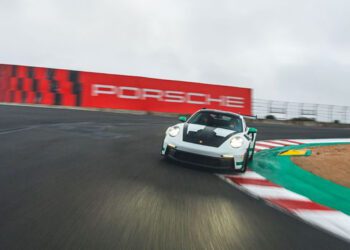 A white Porsche Official Car takes a sharp turn on WeatherTech Raceway Laguna Seca, with a red Porsche sign in the background.