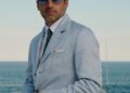 Man in a light blue suit and sunglasses stands outdoors by the ocean, sporting a TAG Heuer Monaco Watches timepiece, with a clear sky and water in the background.