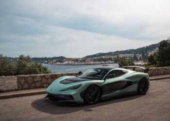 A green and black Rimac Nevera sports car is parked on a coastal road with a stone wall, overlooking the sea and a town with hills in the background, evoking the glamour of the Monaco Grand Prix.