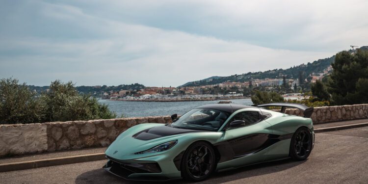 A green and black Rimac Nevera sports car is parked on a coastal road with a stone wall, overlooking the sea and a town with hills in the background, evoking the glamour of the Monaco Grand Prix.
