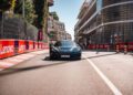 A Rimac Nevera R speeds down a city street lined with Lenovo advertising banners, bordered by modern buildings and a wire fence, evoking the thrill of the Monaco Grand Prix.