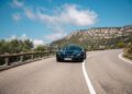 A Rimac Nevera R speeds along a winding mountain road under a partly cloudy blue sky, with rocky hills reminiscent of the Monaco Grand Prix landscape in the background.