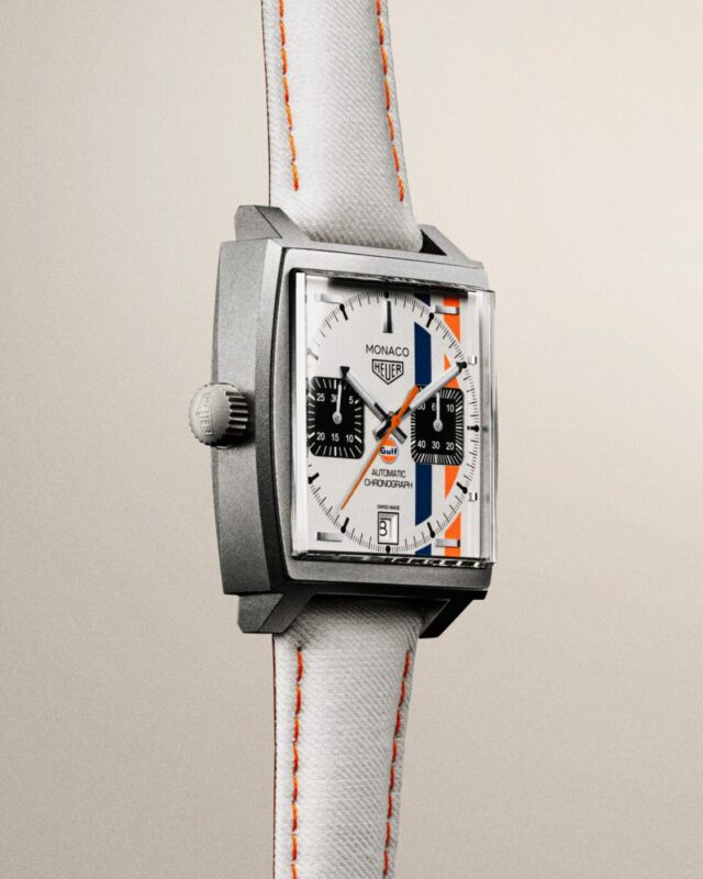 A square-faced TAG Heuer Monaco wristwatch with a white dial, black subdials, orange accents, and a white strap with orange stitching—an iconic tribute to the Monaco Grand Prix.