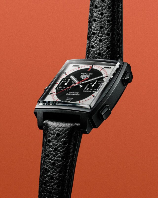 A black TAG Heuer Monaco Grand Prix wristwatch with a square case, black dial, and textured leather strap is displayed against an orange background.
