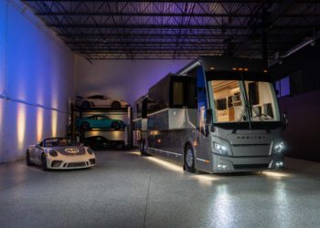 A luxury Off-Grid Prevost Motorcoach by LOKI Coach is parked indoors beside a white Porsche sports car, with additional sports cars stored on racks in the background.