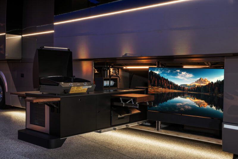 Outdoor kitchen setup under a Prevost off-grid motorcoach featuring a grill, wooden countertops, and a flat-screen TV displaying a scenic landscape.