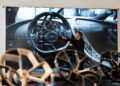A man stands in front of a large screen displaying a detailed image of a Bugatti Tourbillon’s steering wheel and dashboard, pointing toward the controls and showcasing the luxury experience of configuring this masterpiece.