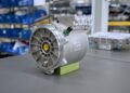 An electric motor with visible wiring sits on a gray workbench in an industrial setting—its innovative design reminiscent of the engineering behind the Bugatti Tourbillon’s Hybrid V16. Shelves and equipment are visible in the background.