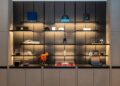 A display shelf in the Washington D.C. showroom with various items, including books, hats, mugs, a bottle, small boxes, and a model helmet, all arranged on illuminated shelves—a true Tourbillon showcase.