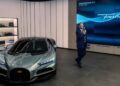 A man in a suit stands beside a silver Bugatti in the Washington D.C. showroom, with a display screen behind him reading "Tourbillon" and "Washington, D.C., 38 May 2023.