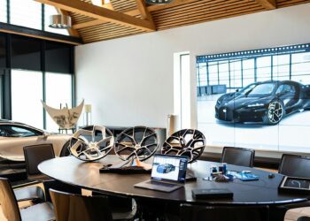 A conference room with car rims on a table, a laptop, and a large screen displaying a Bugatti Tourbillon in the background—perfect for configuring your next luxury experience.