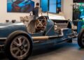 A vintage blue Bugatti race car with exposed engine parts is displayed indoors; two people are seen in the background of a modern Washington D.C. showroom adorned with framed car artwork.