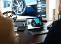 Two people sit at a desk configuring a Bugatti Tourbillon on a laptop, its intricate car wheel design mirrored on the larger screen behind them, highlighting an exclusive luxury experience.