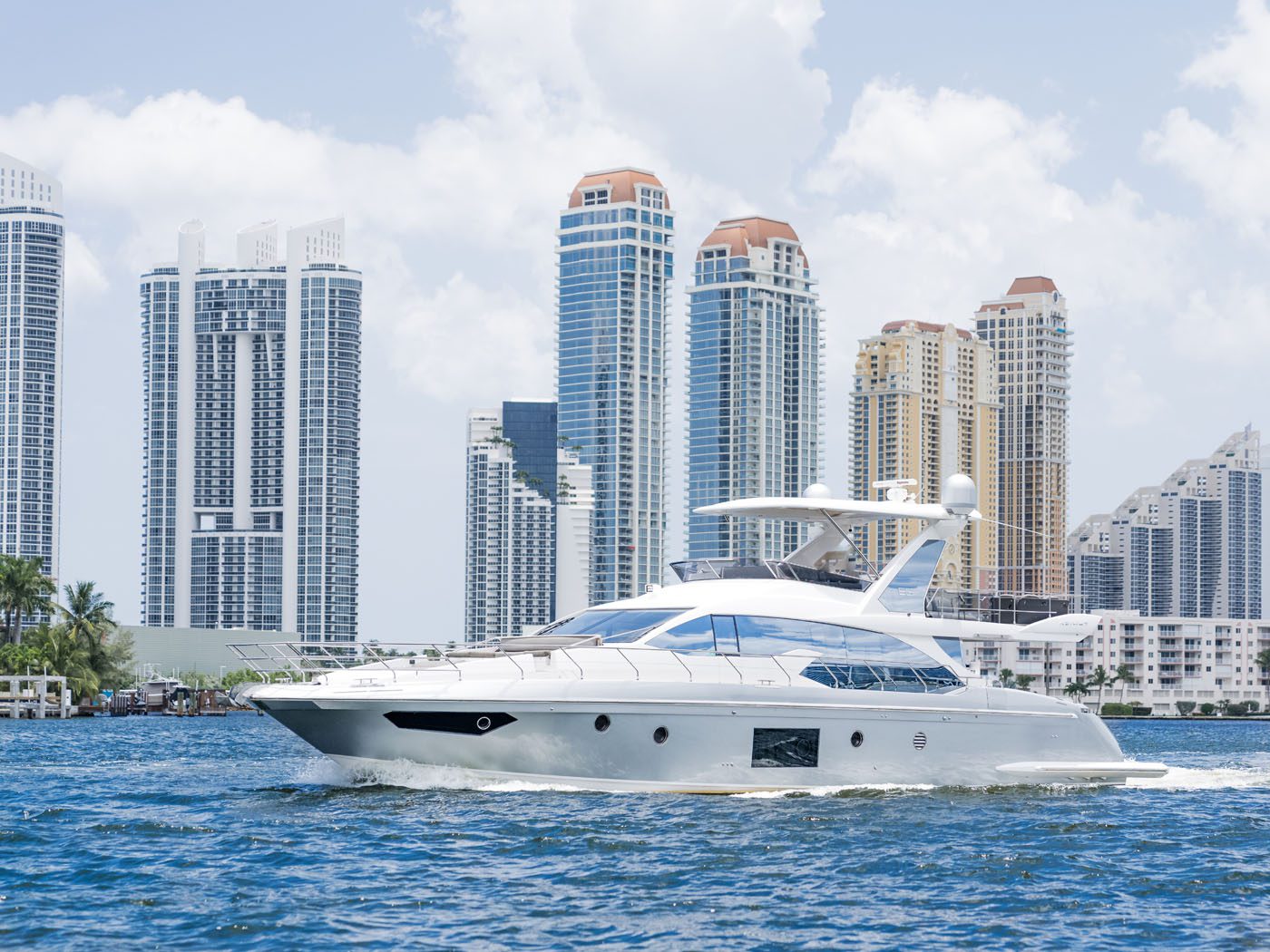 A white luxury yacht with marine protection film sails on blue water, modern high-rise buildings and partly cloudy skies in the background.