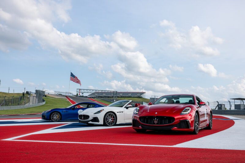 Three track-focused Maserati sports cars—blue, white, and red—are parked on a racetrack at the Circuits of the Americas, with a large American flag in the background under a partly cloudy sky.