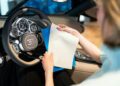 A person sitting in a car holds a blue folder and a white sheet of paper near the steering wheel, which features the Bugatti Tourbillon logo and detailed instrument gauges, capturing an exclusive luxury experience.