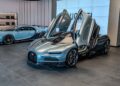 Two silver-blue Bugatti sports cars are displayed in a modern Washington D.C. showroom, with one car's gullwing doors open and the Bugatti logo visible on the wall in the background.