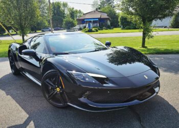 A sleek black Ferrari 296 GTB sports car is parked on a paved driveway, enjoying the Royal Treatment from WrapKings, with trees, grass, and houses in the background on a sunny day.