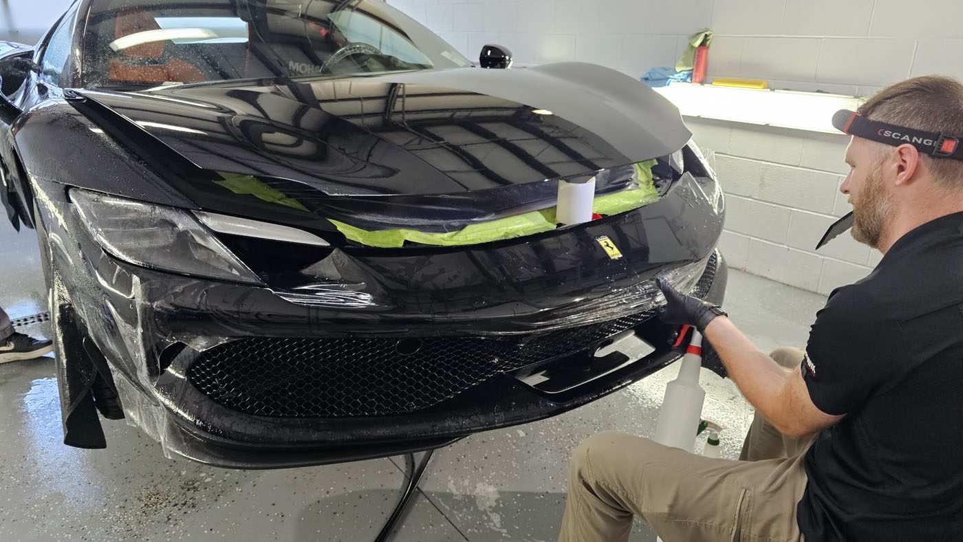 A person gives a Ferrari 296 GTB the Royal Treatment at WrapKings, carefully applying protective film to the front bumper inside a garage.