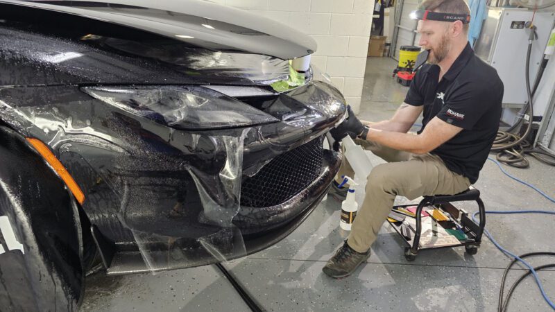At WrapKings, a man applies a protective film to the front bumper of a black Ferrari 296 GTB in an auto workshop, giving it the royal treatment.