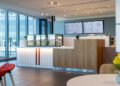 Modern café interior at One Porsche Drive in Atlanta, featuring a wooden counter, pastry display case, digital menu boards, red chairs, and tables with yellow flowers. Large windows provide natural light in this Porsche-inspired space.