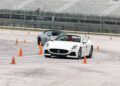 A white Maserati convertible sports car and a silver coupe maneuver through orange traffic cones on the track-focused Circuits of the Americas, with empty grandstands in the background.