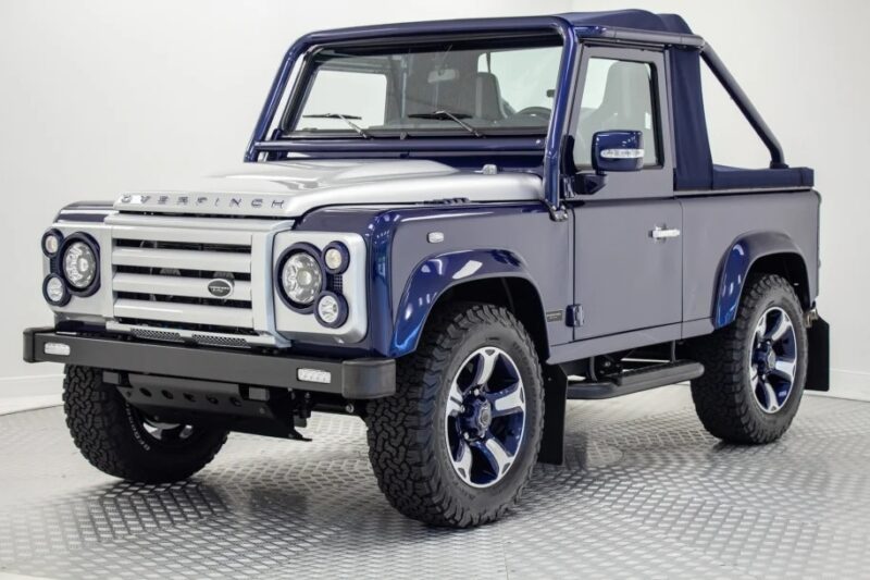 A blue and silver Land Rover Defender Overfinch edition, part of a unique garage, is parked indoors on a checkered floor, viewed from the front left angle.