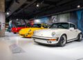 Four classic Porsche sports cars in white, yellow, red, and blue are displayed in a row at One Porsche Drive in Atlanta’s well-lit indoor automobile museum exhibit.