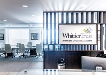 Modern office reception area with a sign reading "Whittier Trust Wealth Management" and a glass-walled conference room with white chairs in the background.