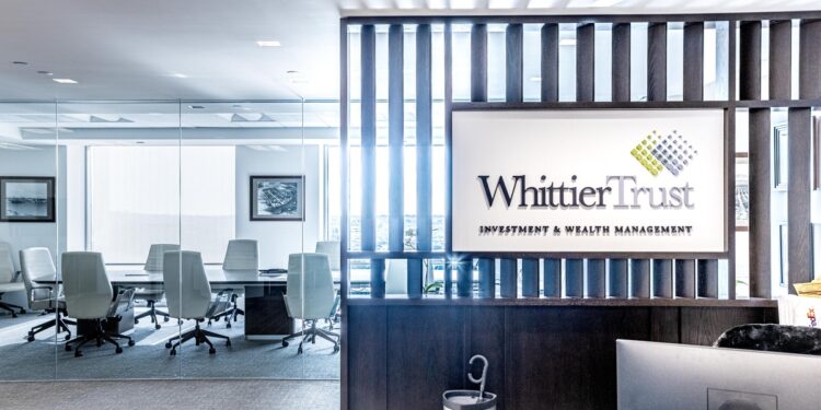Modern office reception area with a sign reading "Whittier Trust Wealth Management" and a glass-walled conference room with white chairs in the background.