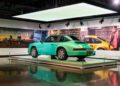 A teal vintage Porsche 911 Carrera is displayed on a raised platform in a modern, well-lit museum at One Porsche Drive in Atlanta, surrounded by striking automotive-themed wall art.