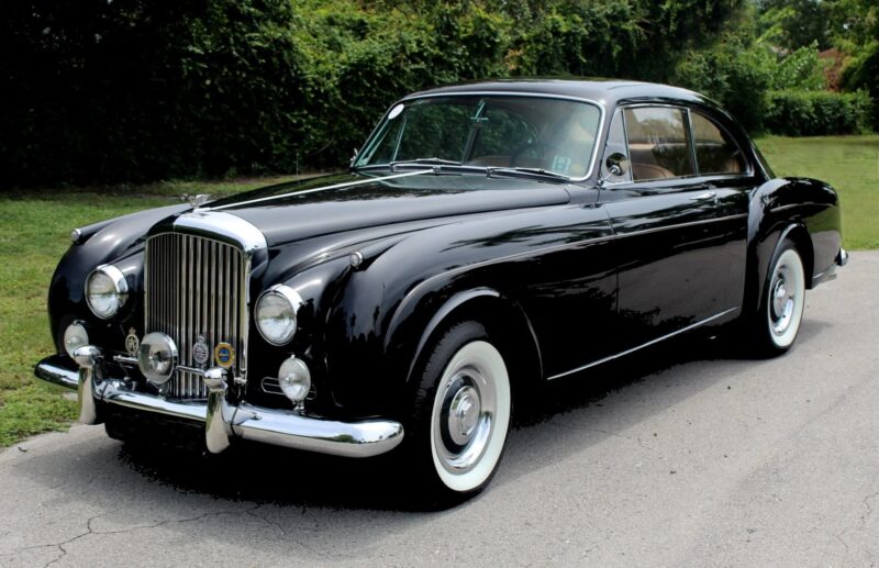 A classic black Bentley coupe with whitewall tires is parked on a paved road, surrounded by greenery—a stunning example among the best Bentleys.